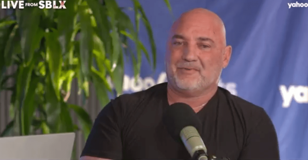 A balding man with a short beard and black shirt sits in front of a microphone, speaking.